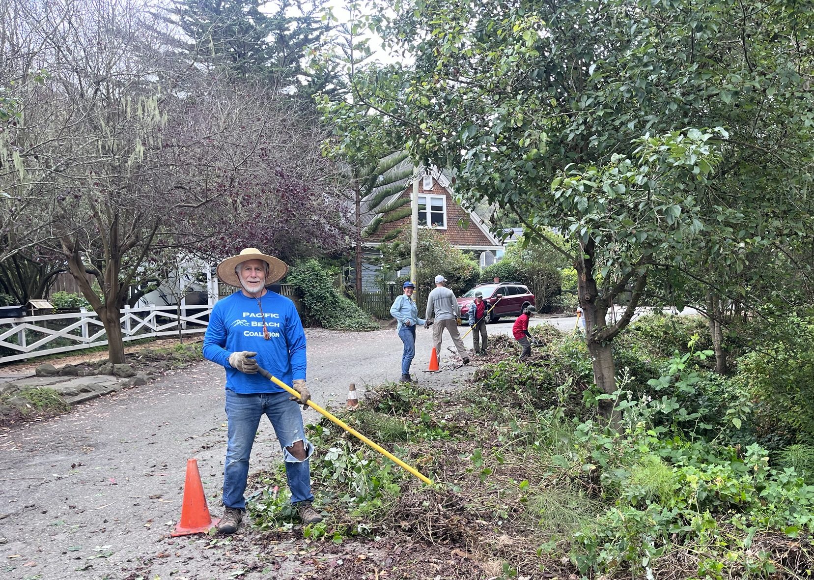 A man with a hat and rake smiles for the camera by a pile of weeds